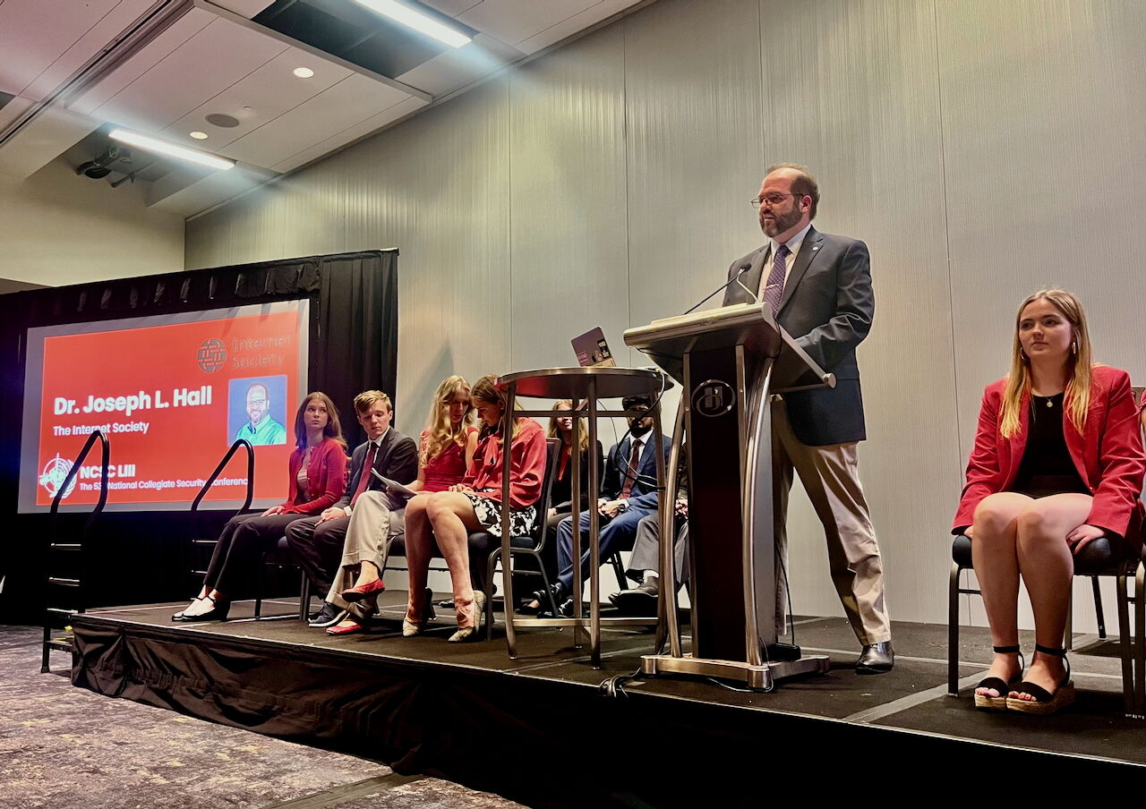 Joseph Lorenzo Hall speaking at a podium with a large projection screen displaying the Internet Society logo in the background.